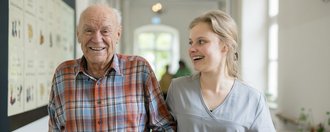 Ein Bewohner und eine Mitarbeiterin gehen eingehakt über einen Flur.