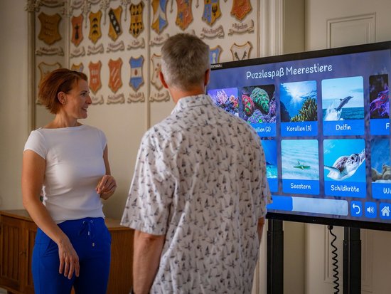 Eine Frau und ein Mann stehen vor einem großen Touchscreen. Auf dem Display läuft das Spiel „Puzzlespaß Meerestiere“ mit Bildern von Korallen, einem Delfin, einem Seestern und einer Schildkröte. Die Frau lächelt den Mann an, der dem Bildschirm zugewandt steht.