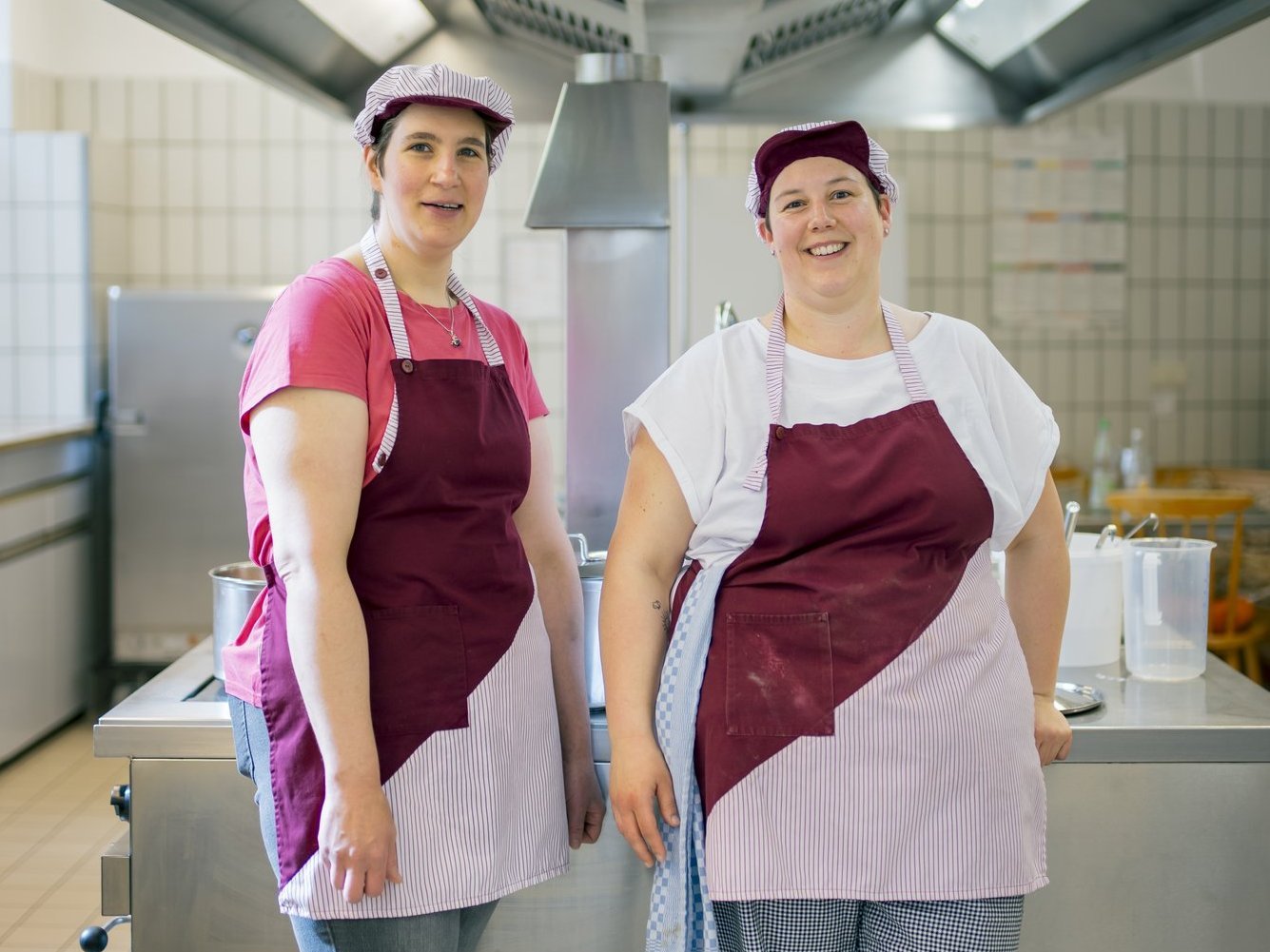 Zwei Mitarbeiterinnen der Hauswirtschaft stehen in der Küche und lachen in die Kamera.