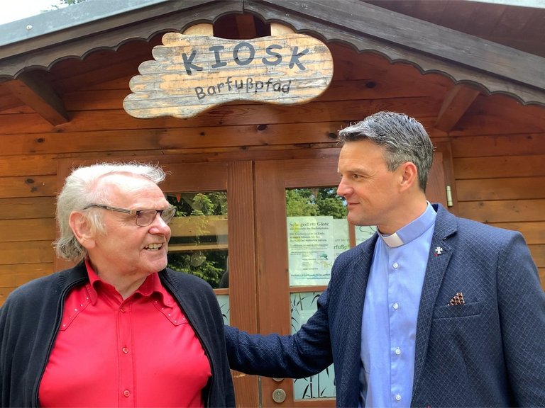 Pater Jörg vor dem Kiosk "Barfußpfad" hört einem älteren Mann zu