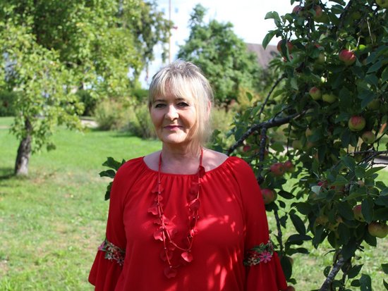 Eine lächelnde Frau mit blonden Haaren steht im Freien neben einem Apfelbaum. Sie trägt ein rotes Oberteil mit floralen Details an den Ärmeln und eine passende Halskette. Im Hintergrund sind eine grüne Wiese und weitere Bäume zu sehen.