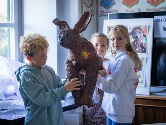 Drei Kinder halten gemeinsam eine große, braune Hasenskulptur. Ein Junge und ein Mädchen tragen das Kunstwerk, während ein drittes Kind im Hintergrund lächelnd zusieht. Es handelt sich um ein kreatives Projekt in einem hellen Raum.
