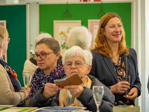 Frauen bei einer Vernissage. Eine Frau mit Brille betrachtet eine Karte, während eine andere lächelnd ein Glas hält. Im Hintergrund hängen gerahmte Bilder an grünen Wänden.