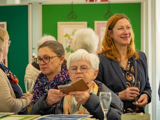 Frauen bei einer Vernissage. Eine Frau mit Brille betrachtet eine Karte, während eine andere lächelnd ein Glas hält. Im Hintergrund hängen gerahmte Bilder an grünen Wänden.
